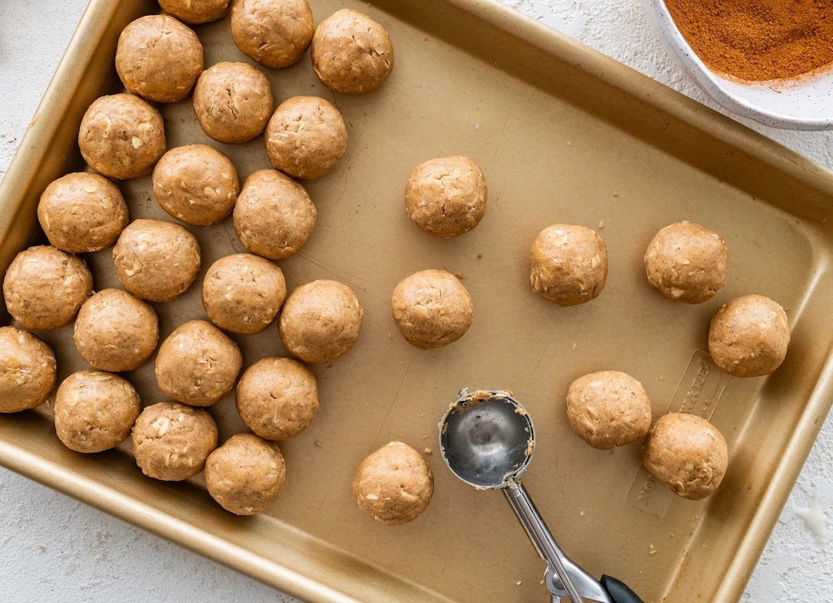 Uncoated protein balls arranged on a baking sheet beside a small cookie scoop.