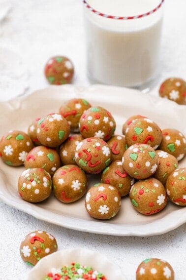 A cream-colored plate piled with sugar cookie protein balls decorated with festive sprinkles, with a glass of milk in the background.