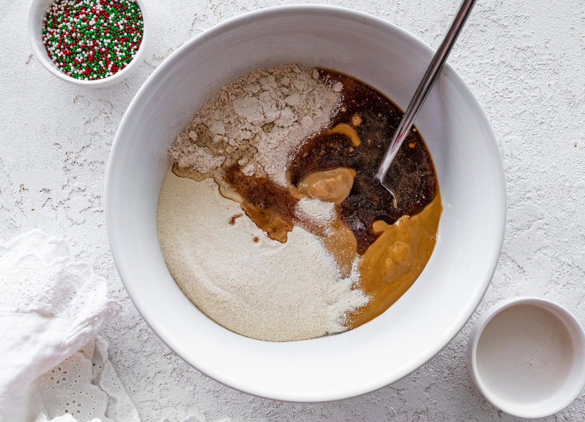 A white mixing bowl showing the unmixed ingredients for sugar cookie protein balls, including oat flour, peanut butter, maple syrup, and protein powder.