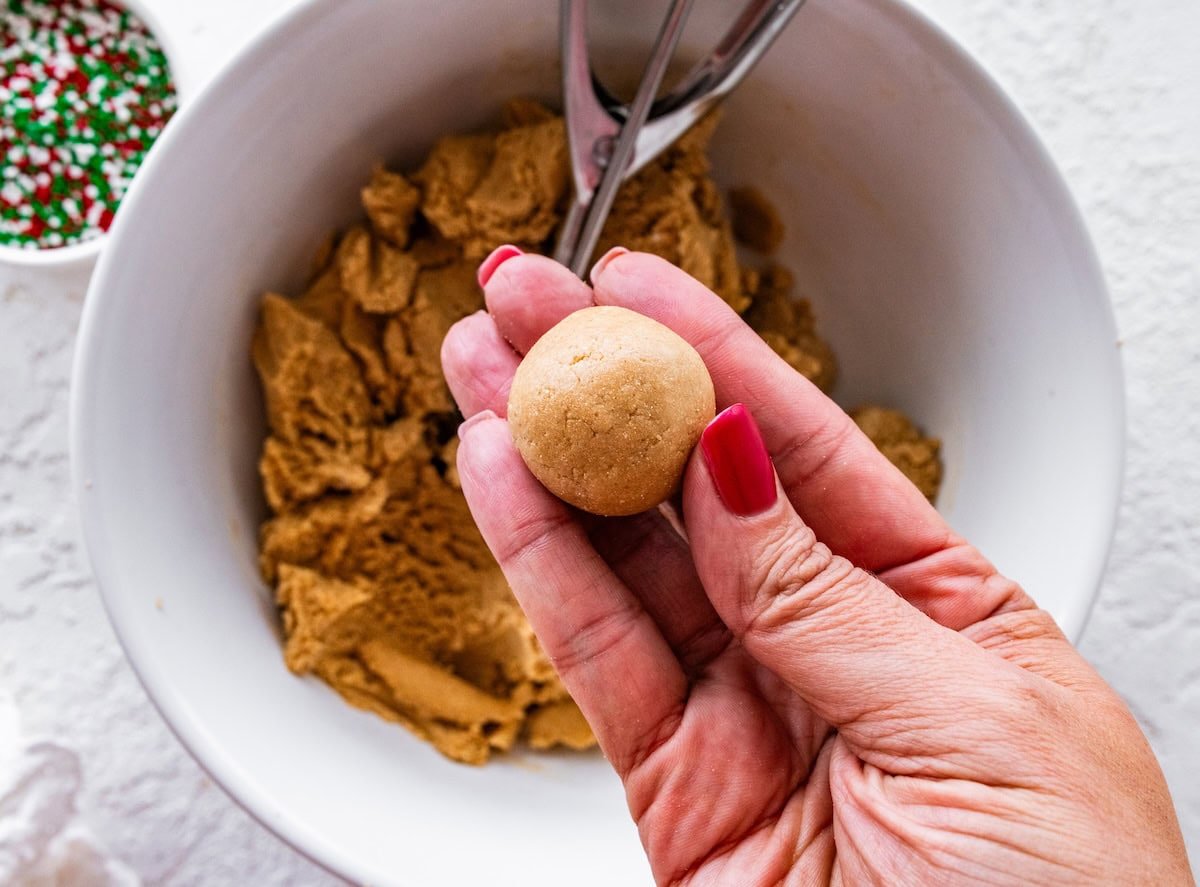 A hand holding a small, round piece of sugar cookie protein ball dough over a bowl of dough, just before rolling in sprinkles.