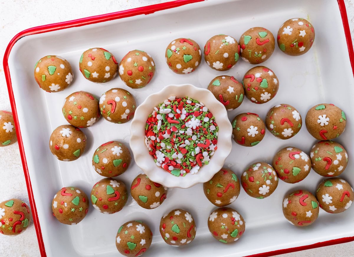 A rectangular tray lined with sugar cookie protein balls surrounding a small dish of red, white, and green sprinkles in the center.