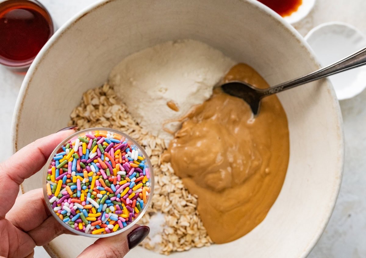 A small bowl of sprinkles being held over a large mixing bowl with cashew butter, protein powder, and rolled oats.