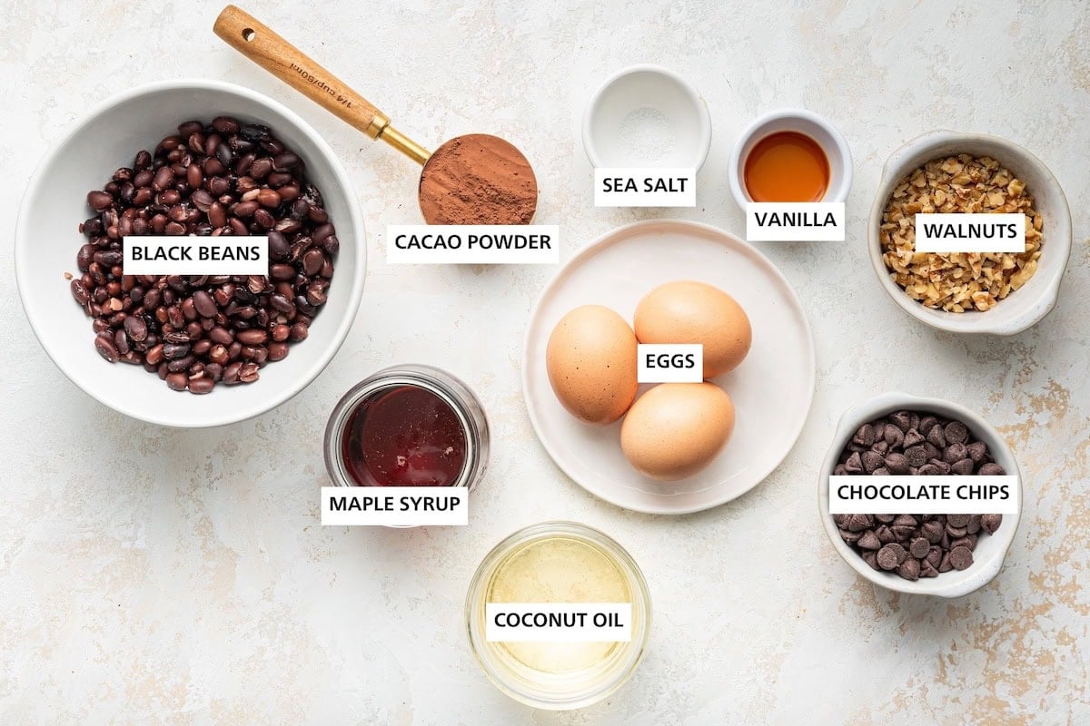 Ingredients measured out to make Fudgy Black Bean Brownies: black beans, maple syrup, cacao powder, coconut oil, eggs, sea salt, vanilla, chocolate chips and walnuts.