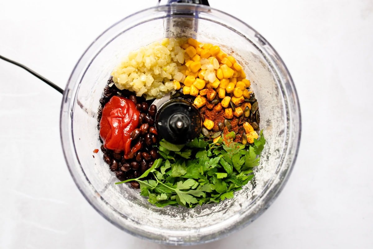 Corn, black beans, ketchup, parsley, pepitas, garlic, onion, and spices in a food processor used for black bean burgers.