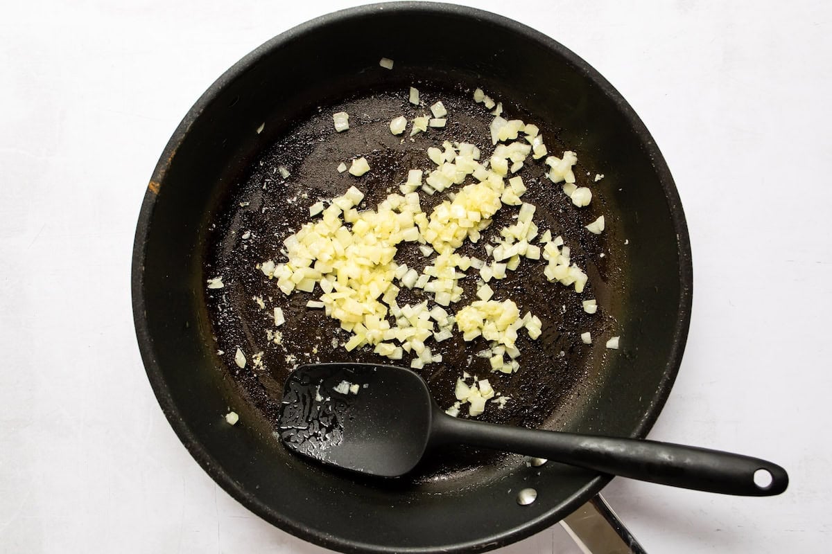 Garlic and diced onion being sauteed in a black skillet with a silicon spatula.