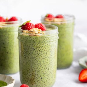 Jars of creamy matcha chia pudding topped with fresh strawberries on a white surface.