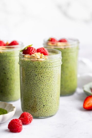 Jars of creamy matcha chia pudding topped with fresh strawberries on a white surface.