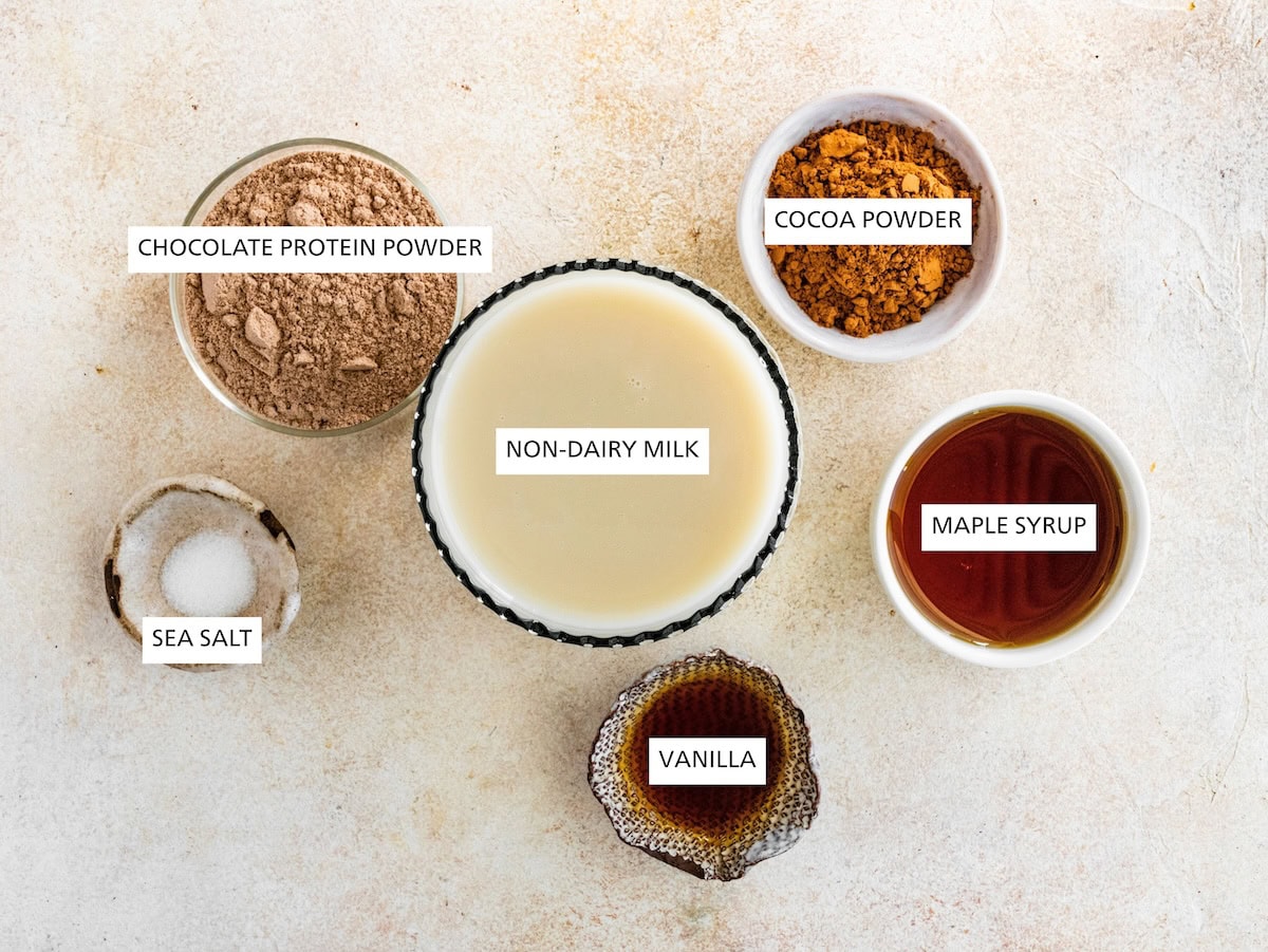 Ingredients measured out to make Protein Hot Chocolate: cocoa powder, maple syrup, vanilla, oat milk, chocolate protein powder and sea salt.