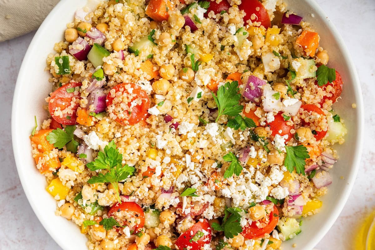 A mixed quinoa chickpea salad with tomatoes, cucumbers, feta, and parsley, in a white bowl.