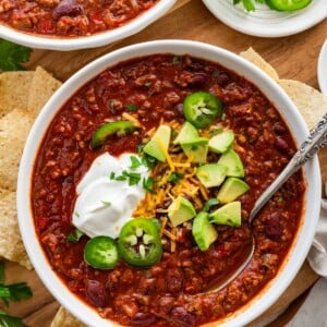 A bowl of slow cooked bean and beef chili topped with avocado, cheese, sour cream, and jalapeno slices.