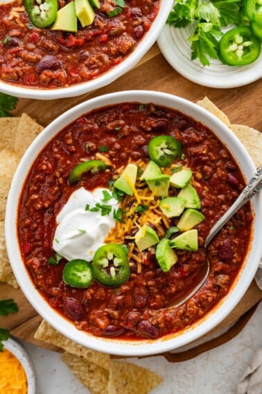 A bowl of slow cooked bean and beef chili topped with avocado, cheese, sour cream, and jalapeno slices.