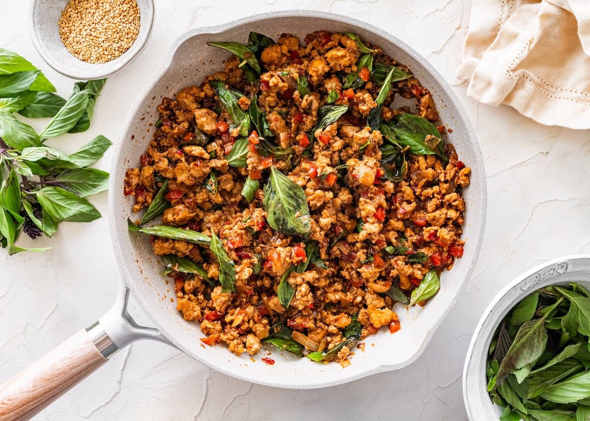 Saucy Thai basil chicken cooking in a skillet with fresh basil leaves stirred in.