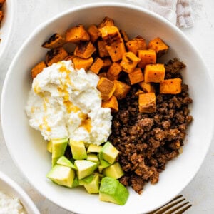 Ground beef in a white bowl with roasted sweet potatoes, cottage cheese, avocado chunks and a drizzle of hot honey.