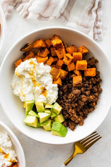 Ground beef in a white bowl with roasted sweet potatoes, cottage cheese, avocado chunks and a drizzle of hot honey.