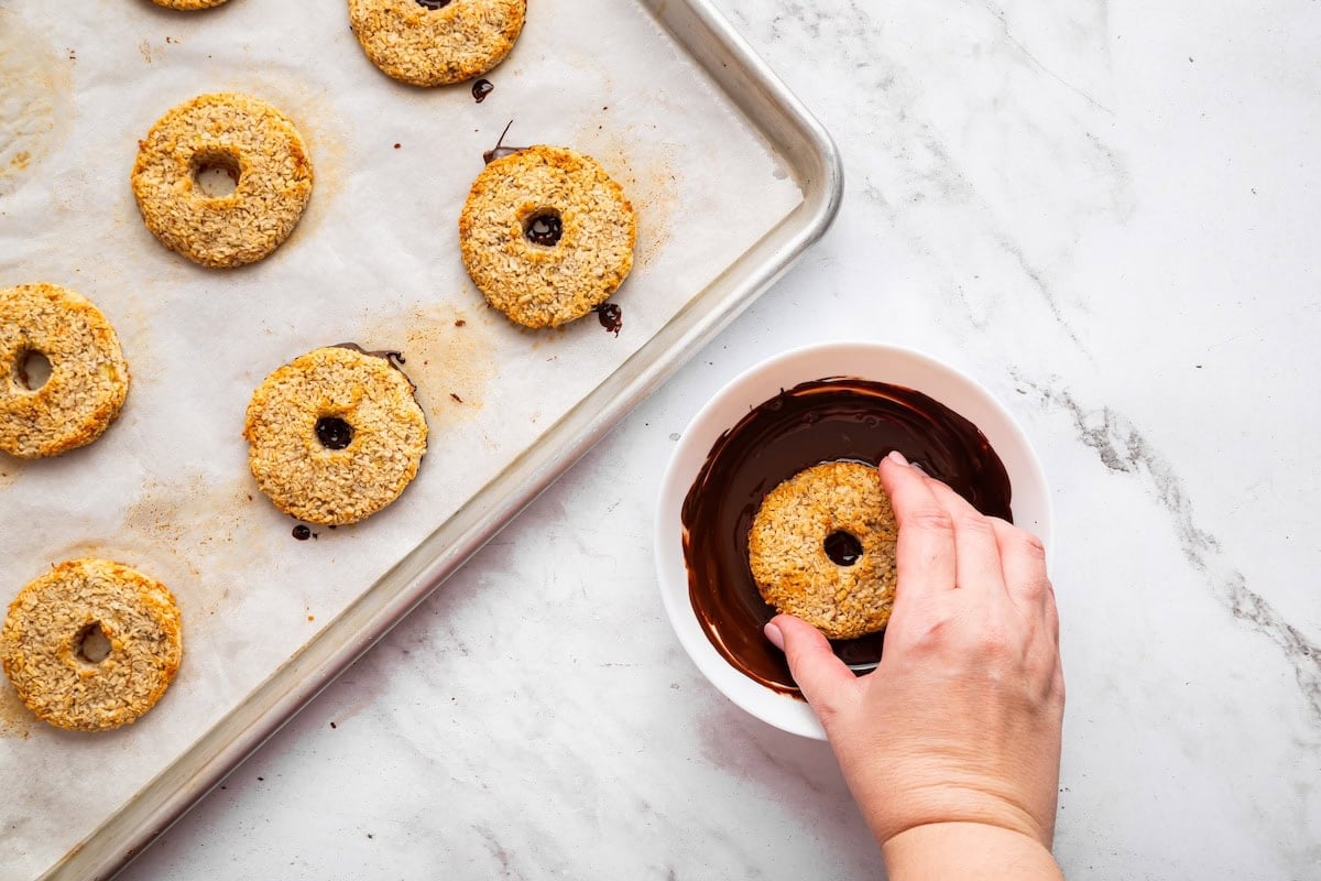 A womans hand dips a banana samoas cookie into a small bowl of melted chocolate just to coat the bottom of the cookie.