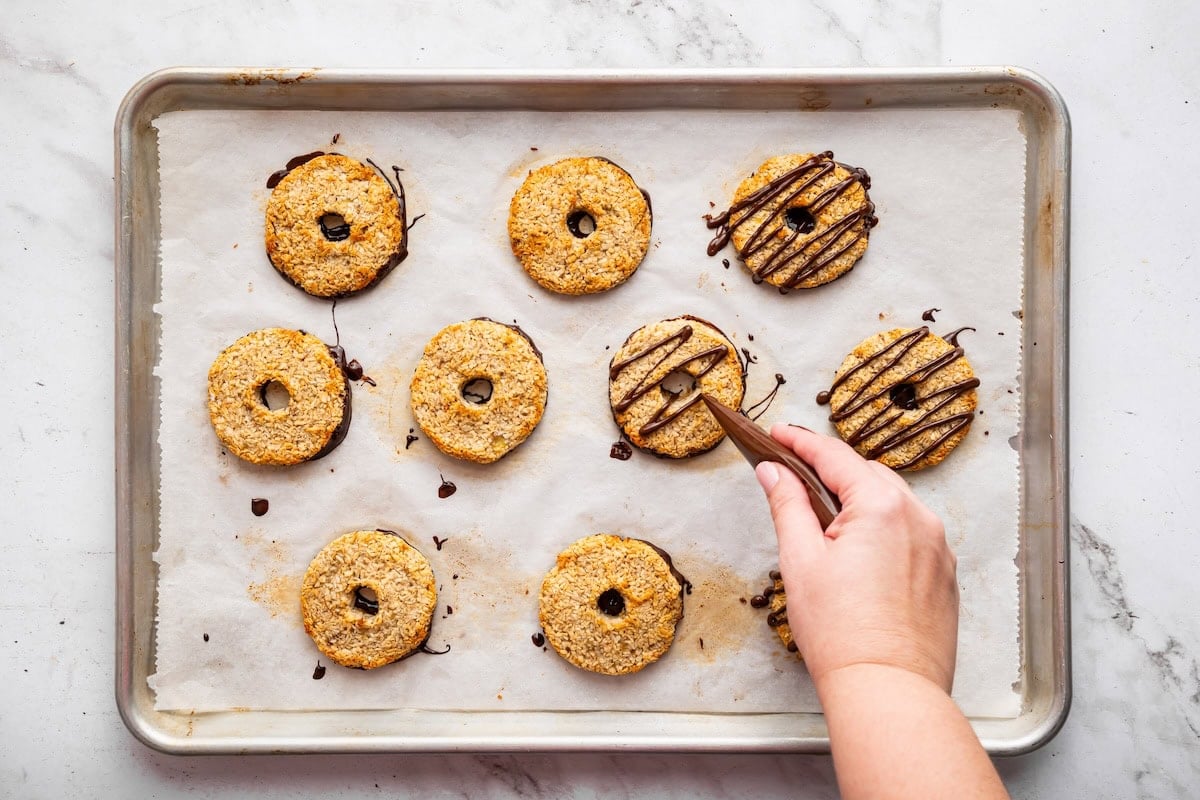 A womans hand uses a small pipping bag to drizzle chocolate on top of the banana samoas cookies on a baking tray.