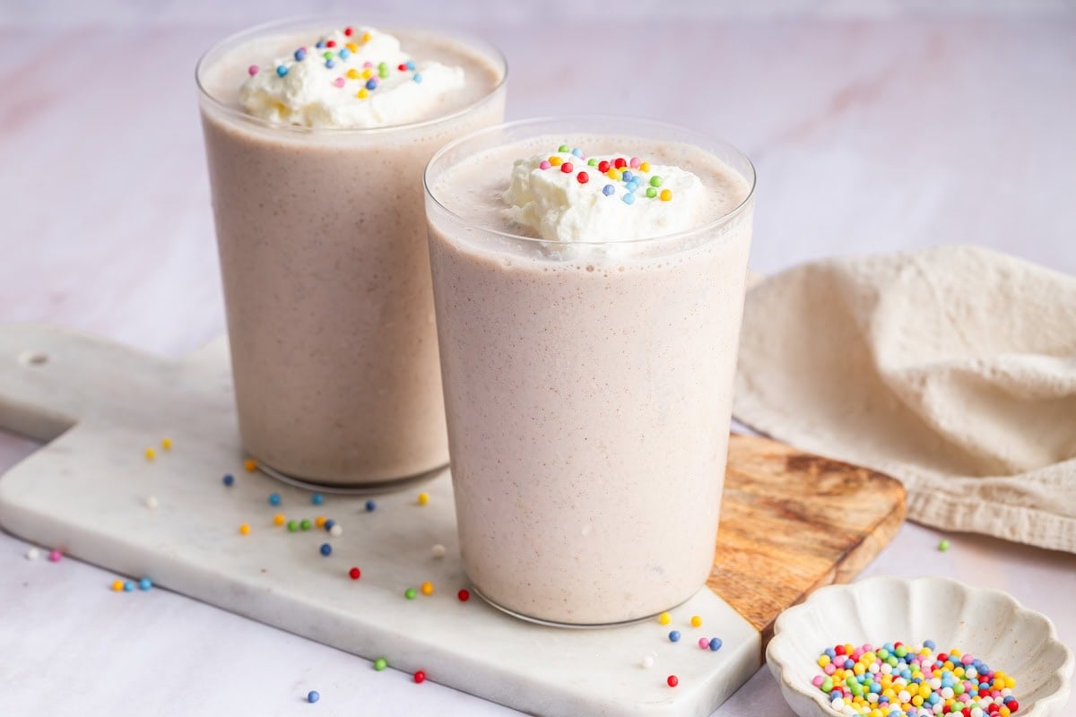 Two cake batter protein shakes in glasses topped with whipped cream and rainbow sprinkles.