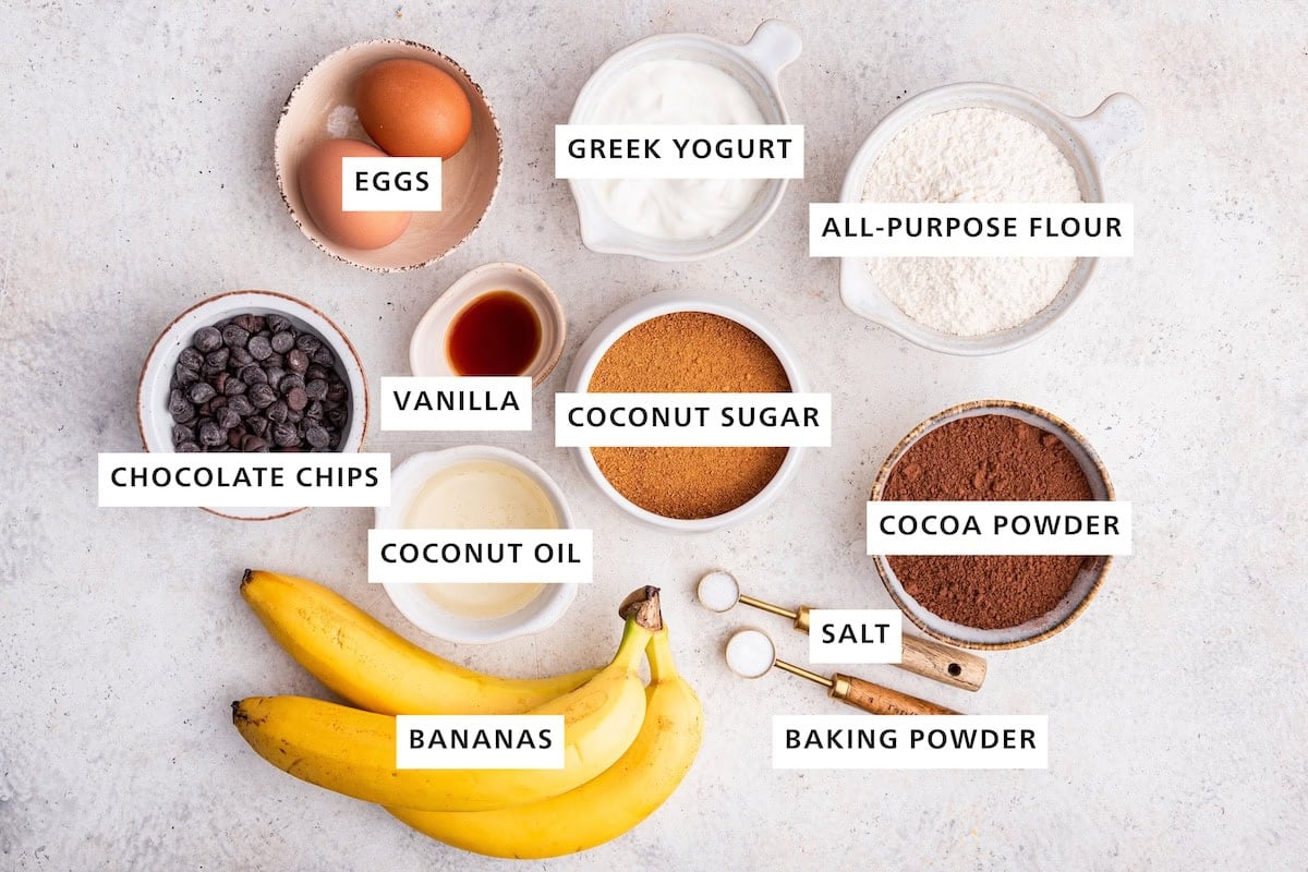 Ingredients measured out to make Chocolate Banana Bread: flour, cocoa powder, baking soda, coconut sugar, sea salt, Greek yogurt, eggs, vanilla, coconut oil, bananas and chocolate chips.