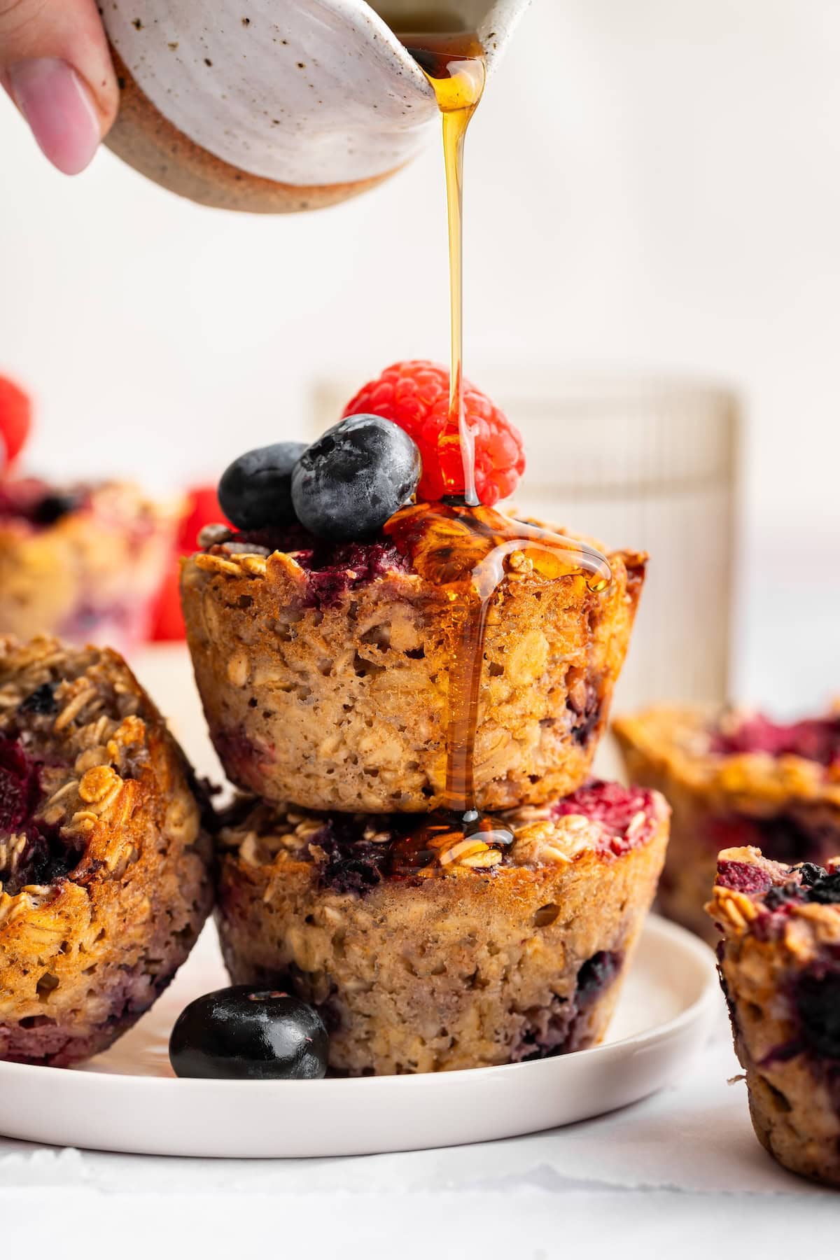 Two cottage cheese baked oatmeal cups stacked on top of each other topped with fresh blueberries and raspberries. A woman's hand is drizzling maple syrup on top.