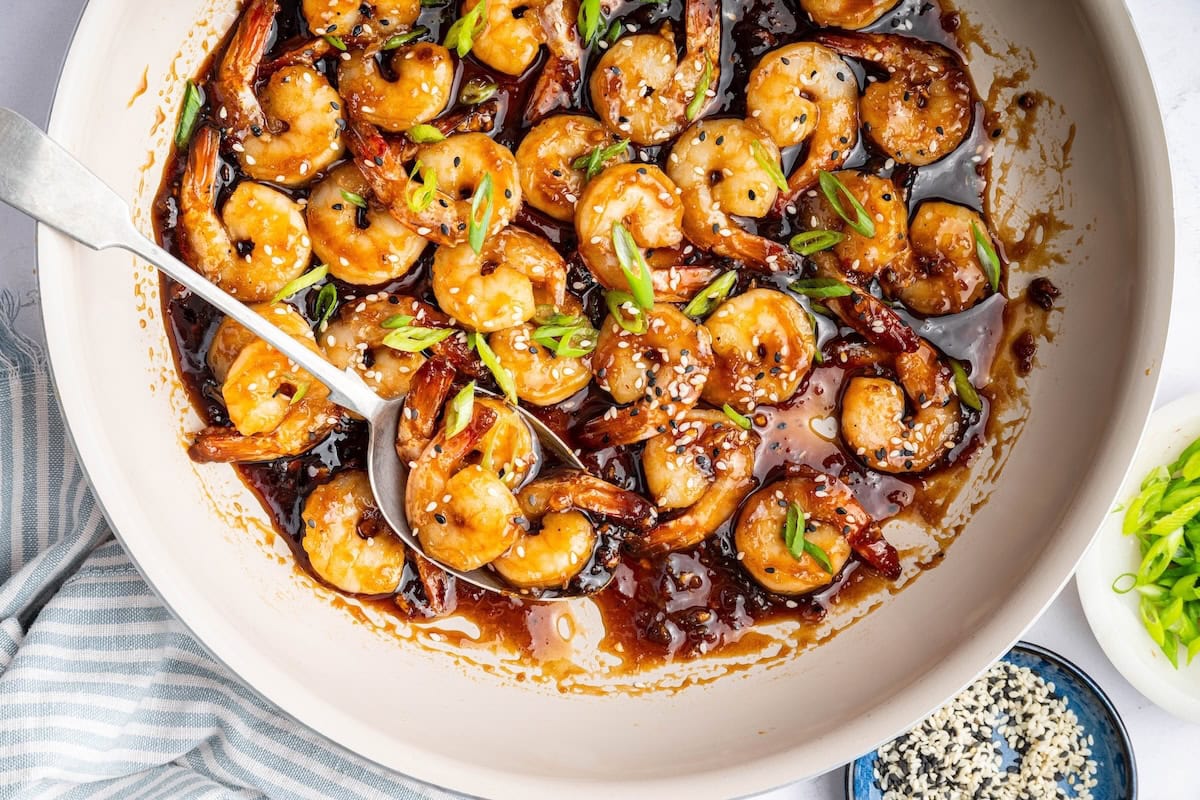 Shrimp coated in a honey garlic sauce in a skillet. There is a silver spoon in the skillet and the shrimp is topped with sesame seeds and green onion slices.