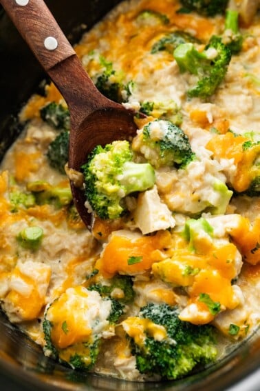 Chicken, rice, cheddar cheese, and broccoli in a slow cooker with a wooden serving spoon.