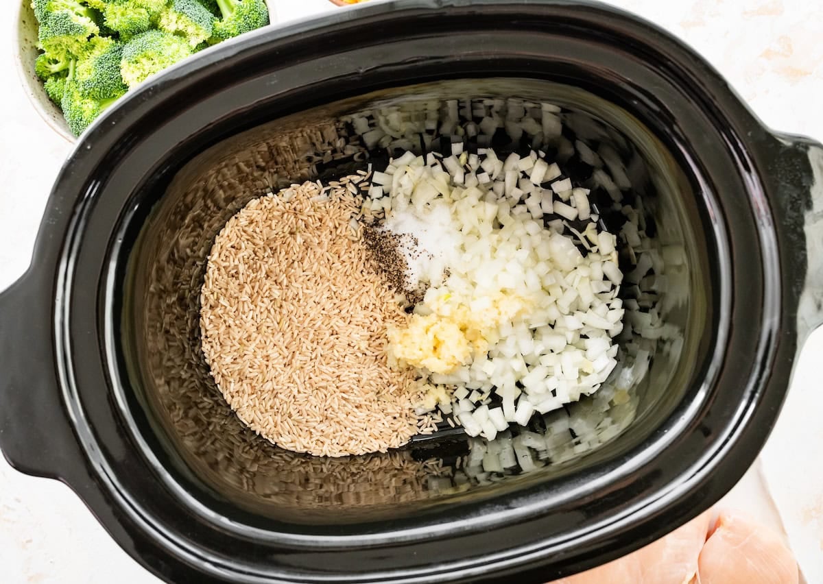Diced onion, minced garlic, brown rice, ground black pepper, and salt in a slow cooker before being cooked.