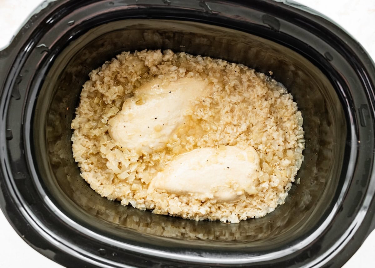 Cooked chicken breasts in a slow cooker with chicken broth, diced onion, minced garlic, brown rice, ground black pepper, and salt.