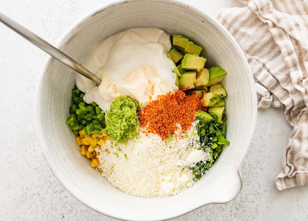 A large mixing bowl with mayo, tajin, lime zest, cojita cheese, corn, avocado, and jalapeno.