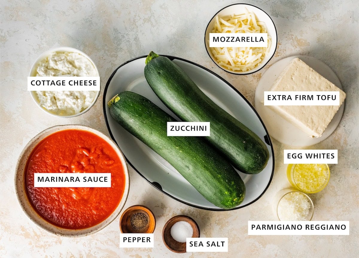 Ingredients measured out to make Tofu Lasagna with Zucchini Noodles: marinara sauce, cottage cheese, sea salt, pepper, zucchini, parmigiano reggiano, extra firm tofu, egg whites and mozzarella.
