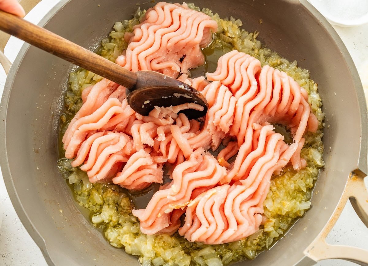 Raw ground turkey being broken up with a wooden spoon in a skillet with sauteed onion and garlic.