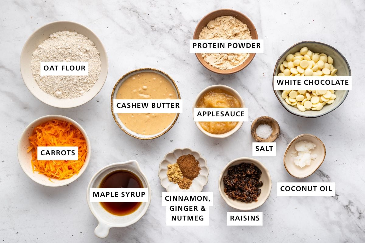 Ingredients for the carrot cake protein balls: oat flour, carrots, cashew butter, applesauce, maple syrup, cinnamon, ginger, nutmeg, raisins, salt, coconut oil, white chocolate and protein powder.