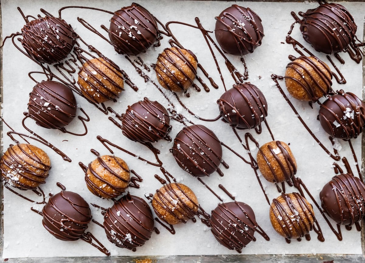 Banana bread protein balls on parchment paper. Half are drizzled in chocolate and half are dipped fully in chocolate. All are topped with flakey sea salt.