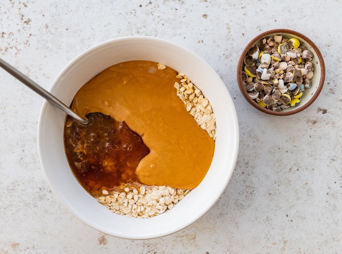 A mixing bowl with quick oats, protein powder, peanut butter, and honey. There is a small bowl of chocolate Cadbury Eggs nearby.