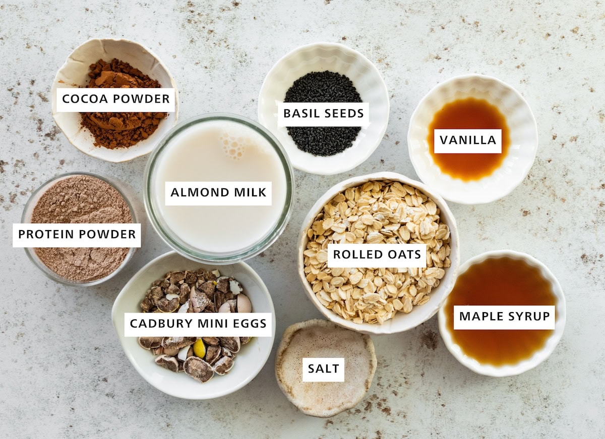 Ingredients measured out for Cadbury Mini Egg overnight oats: almond milk, basil seeds, vanilla, salt, rolled oats, maple syrup, cadbury min ieggs, protein powder, cocoa powder.