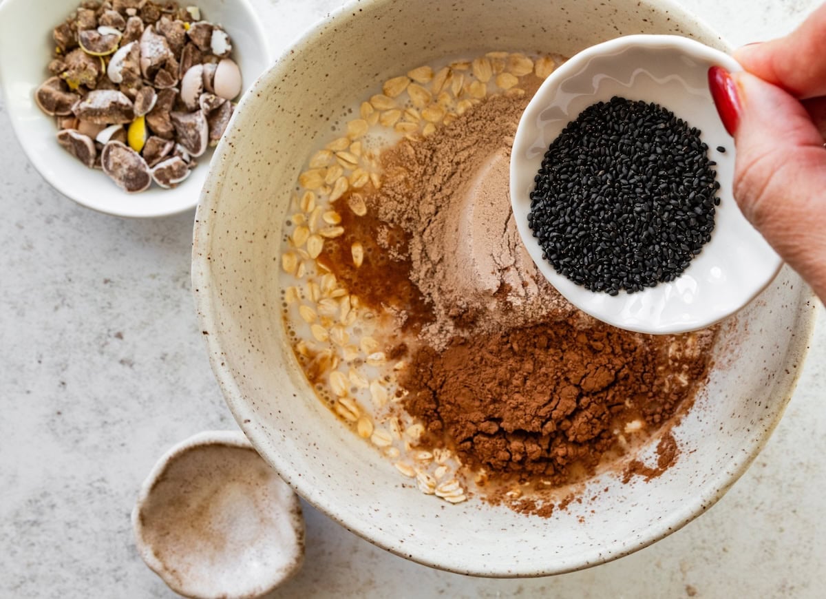 A womans hand holds a small bowl of basil seeds over a large bowl of oats, protein powder, cacao powder, and spices used for overnight oats.
