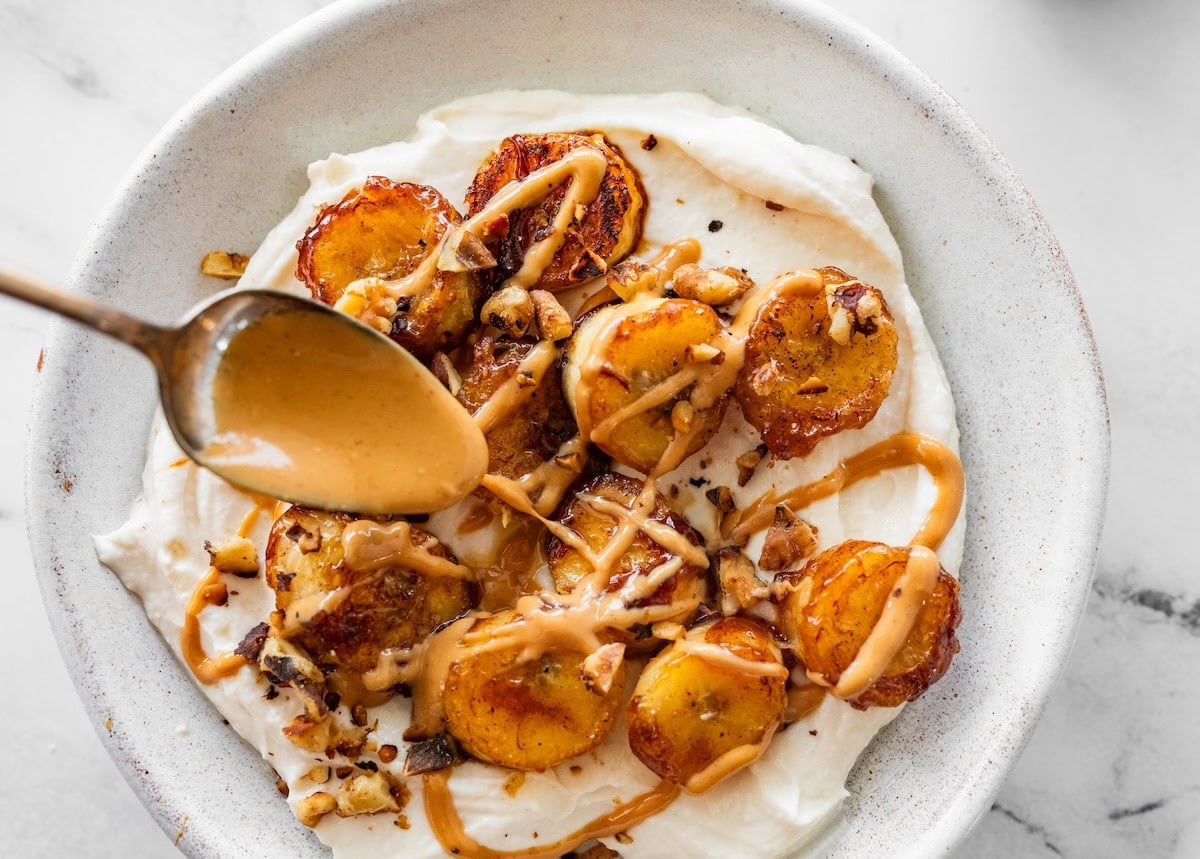 A spoon drizzles peanut butter on caramelized banana slices in a bowl of greek yogurt.