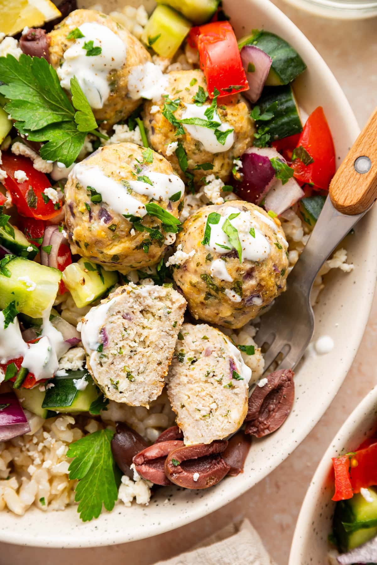 A Greek turkey meatball bowl with brown rice, tzatziki, cucumber tomato salad, olives, and fresh herbs.