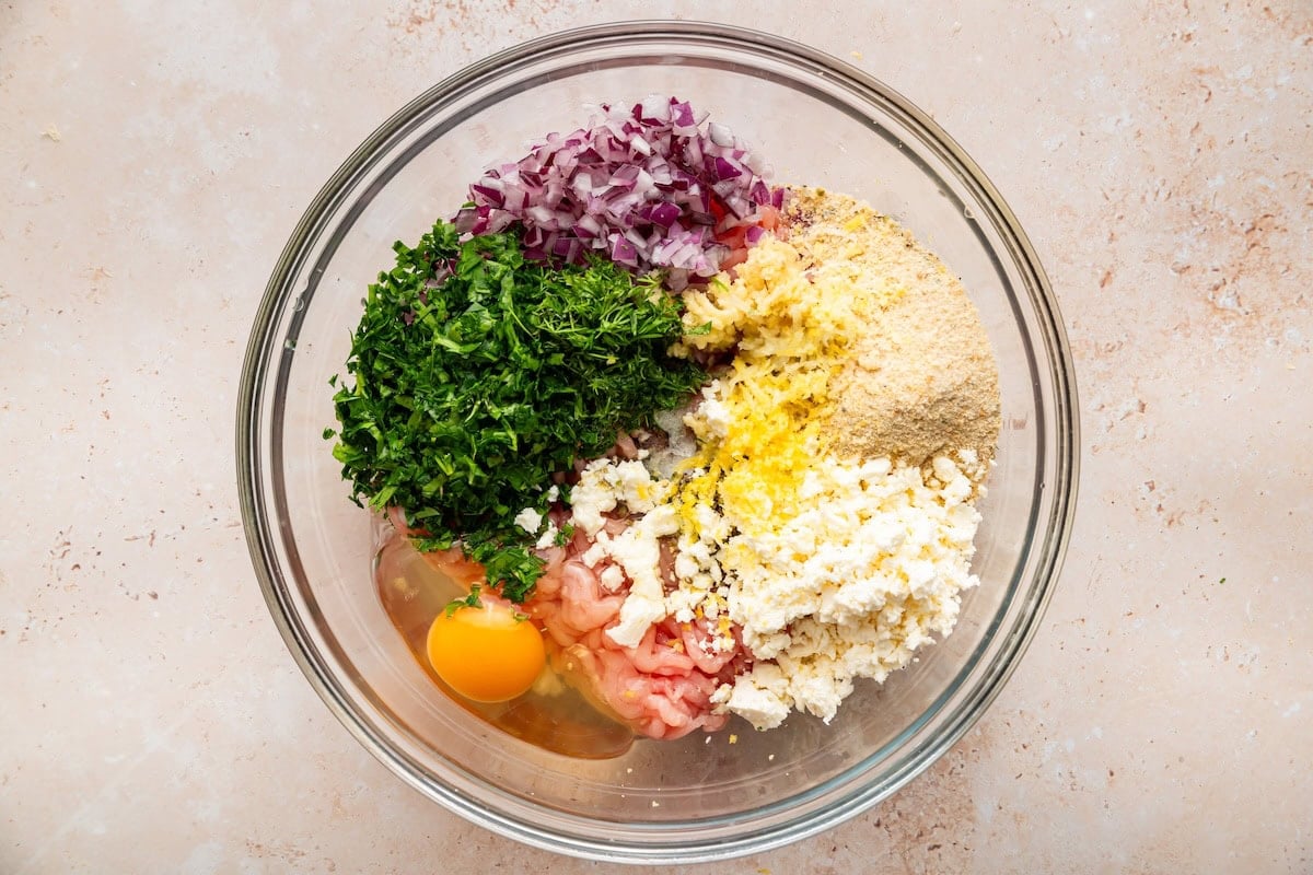 A large glass mixing bowl with ingredients used to make Greek turkey meatballs before being mixed.