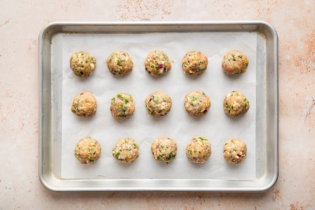 Fifteen Greek turkey meatballs arranged on a metal baking tray before being baked.