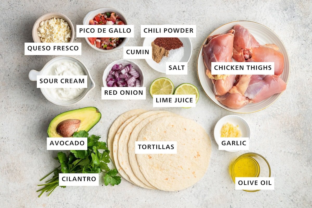 Ingredients measured out to make Healthy Chicken Tacos: chicken thighs, garlic, olive oil, lime, chili powder, salt, cumin, red onion, tortillas, pico de gallo, queso fresco, sour cream, avocado and cilantro.