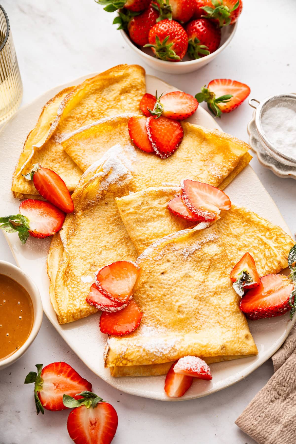 Five cottage cheese protein crepes sprinkled with powdered sugar and topped with sliced strawberries on a plate.