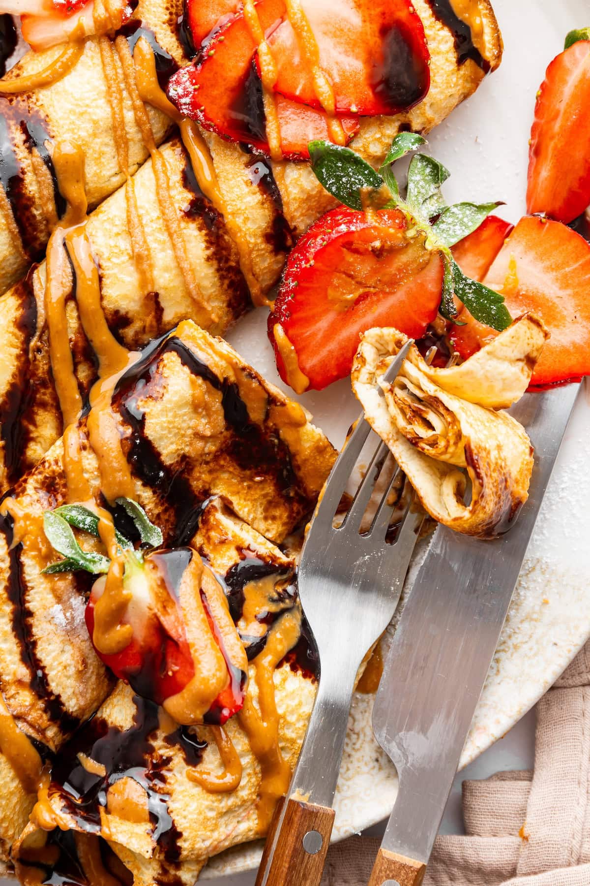 Protein crepes on a plate topped with almond butter, chocolate, and strawberries. A fork and a knife sit on the plate with a bite size piece of the crepe.