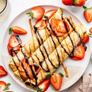 Three protein crepes rolled up on a plate topped with a drizzle of nut butter, chocolate, and a topping of fresh sliced strawberries.