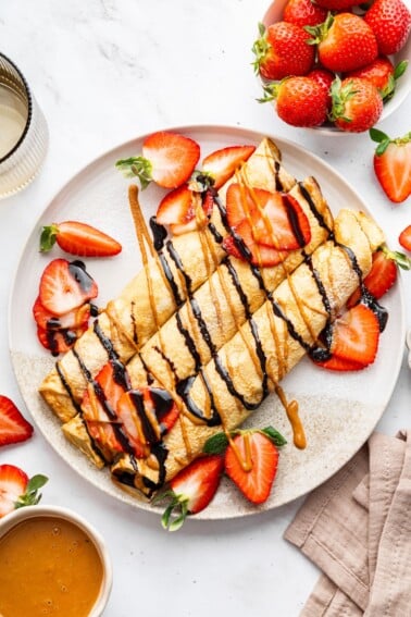 Three protein crepes rolled up on a plate topped with a drizzle of nut butter, chocolate, and a topping of fresh sliced strawberries.