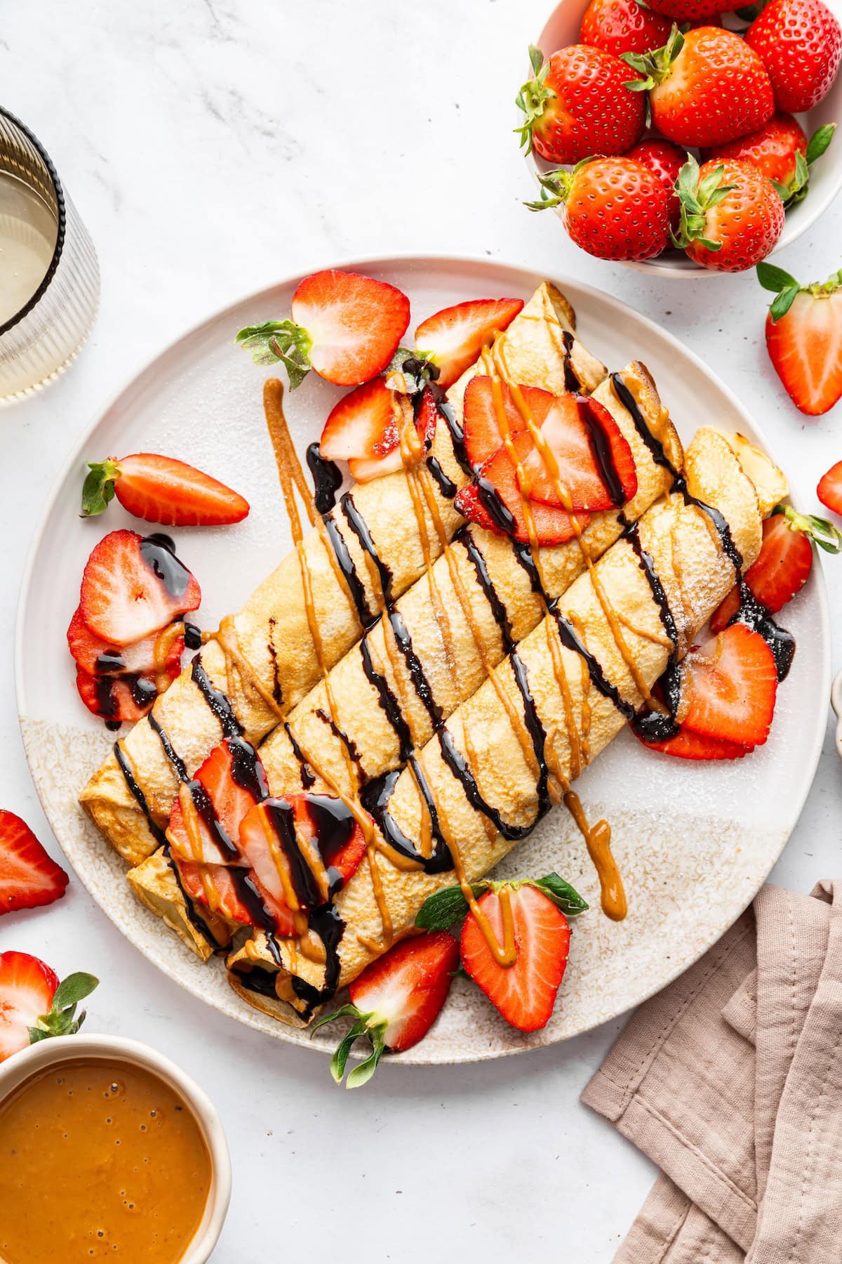 Three protein crepes rolled up on a plate topped with a drizzle of nut butter, chocolate, and a topping of fresh sliced strawberries.