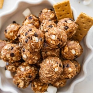 No-bake s&rsquo;mores protein balls served on a decorative white plate alongside graham crackers and mini marshmallows.