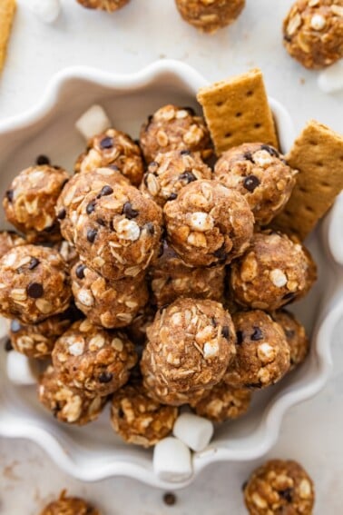 No-bake sโmores protein balls served on a decorative white plate alongside graham crackers and mini marshmallows.