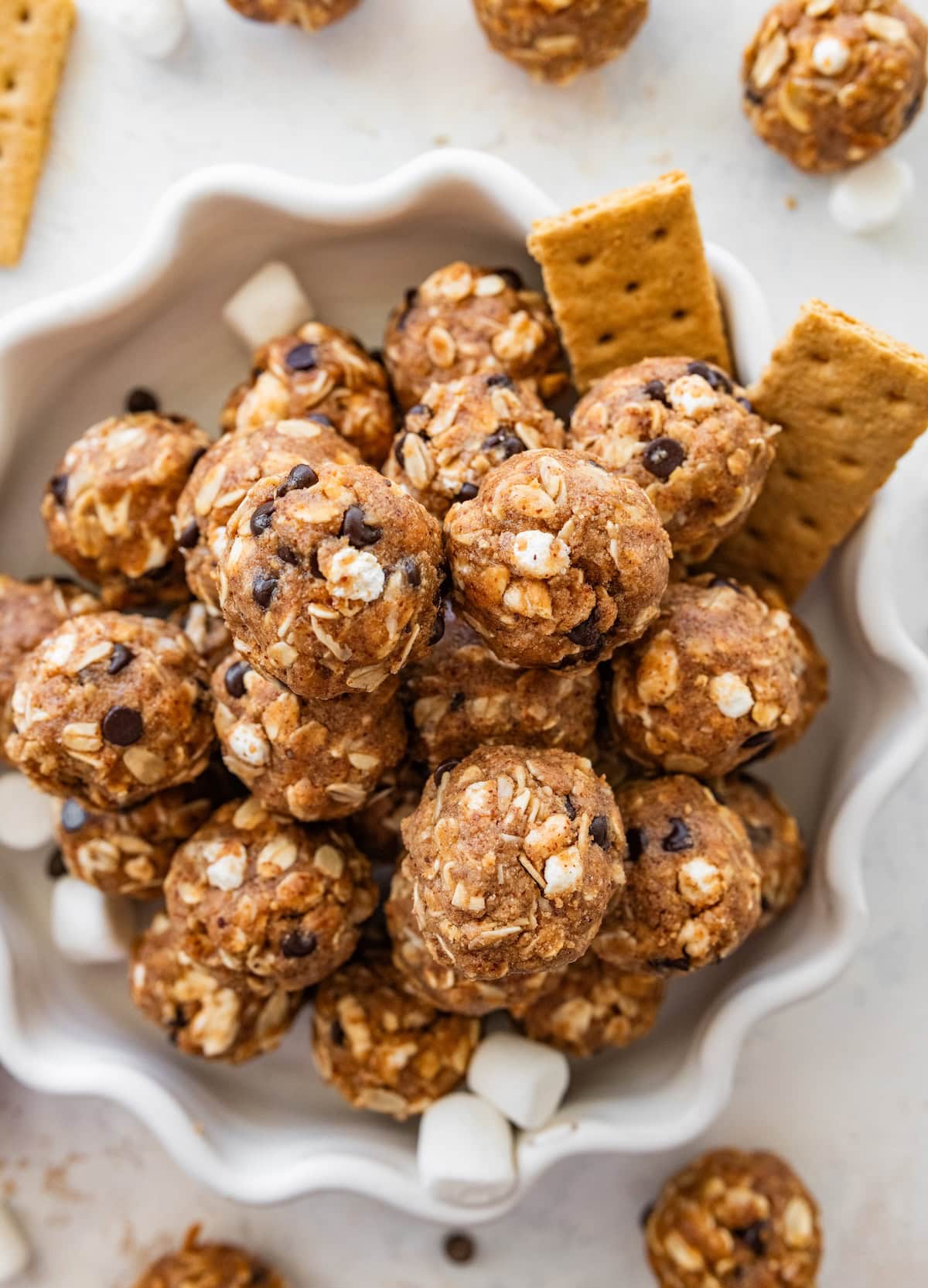 No-bake s&rsquo;mores protein balls served on a decorative white plate alongside graham crackers and mini marshmallows.