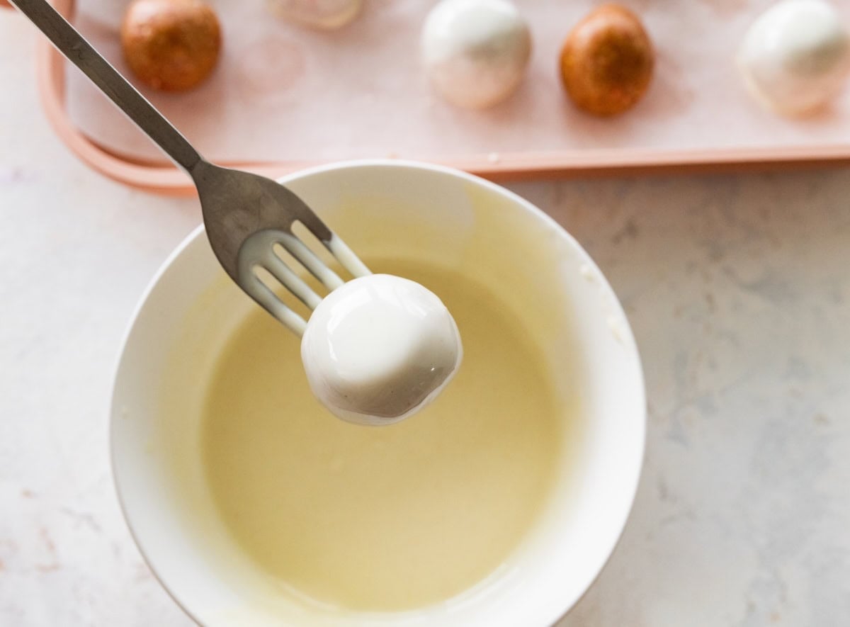 A fork dipping a strawberry protein ball in melted white chocolate.