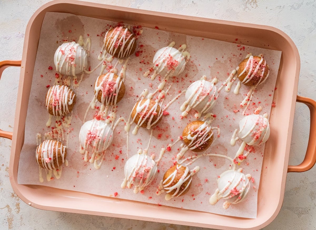 Strawberry protein balls arranged on a tray with white chocolate drizzle and crushed strawberries.
